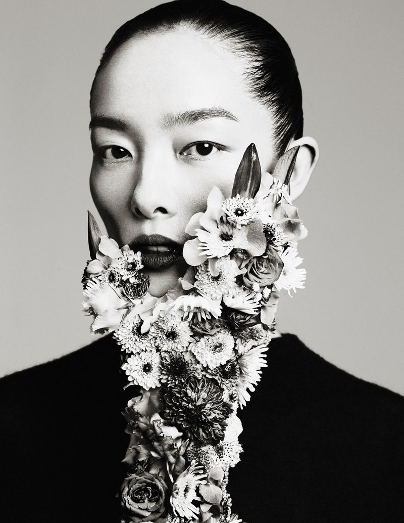 Monochromatic portrait of a serene adult woman adorned with floral beard, exuding a blend of natural artistry against a plain backdrop.