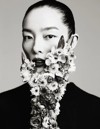 Monochromatic portrait of a serene adult woman adorned with floral beard, exuding a blend of natural artistry against a plain backdrop.