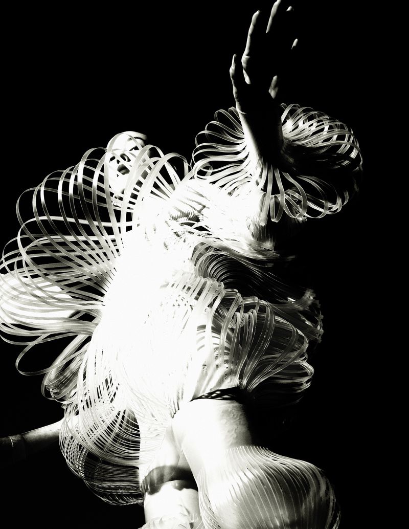 Artistic black and white studio shot of a human figure wearing a translucent sculptural garment made of layered, spiraling elements.