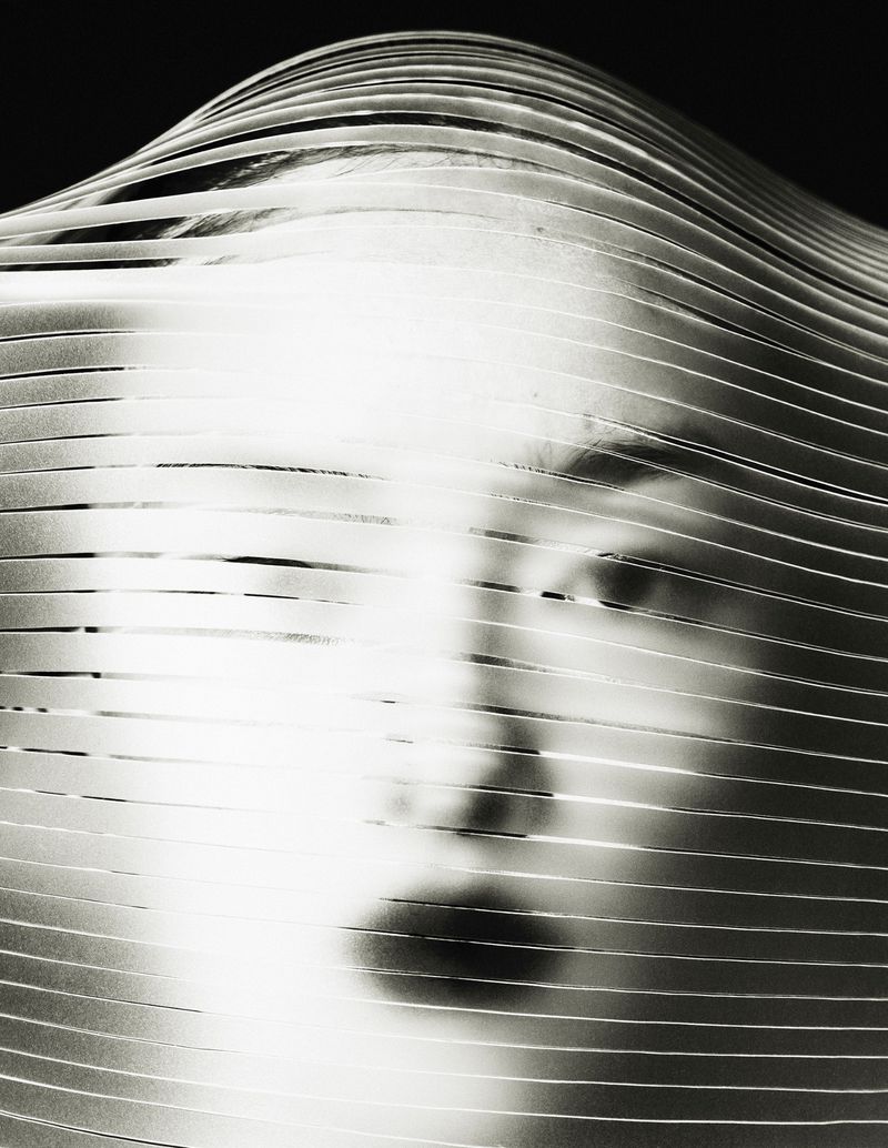 Striking black-and-white portrait of a person's face obscured by parallel ribbons, creating an abstract, textured effect in studio.