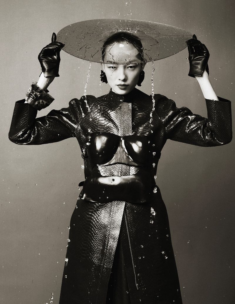 Monochrome portrait of Asian female in glossy black outfit with a clear plastic umbrella, standing against a gray background while water falls.