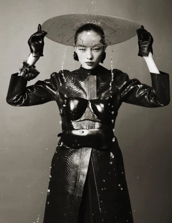 Monochrome portrait of Asian female in glossy black outfit with a clear plastic umbrella, standing against a gray background while water falls.