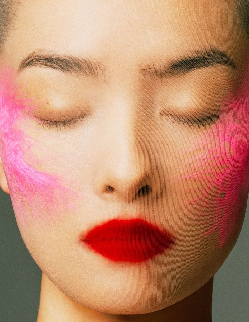 Close-up portrait of a woman with closed eyes, red lipstick, and pink feather-like accents on her cheeks, against a neutral backdrop.