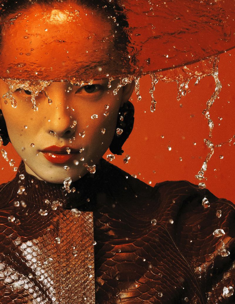 Asian woman in red lipstick and a snakeskin jacket is splashed with water in front of a red background in a fashion portrait.