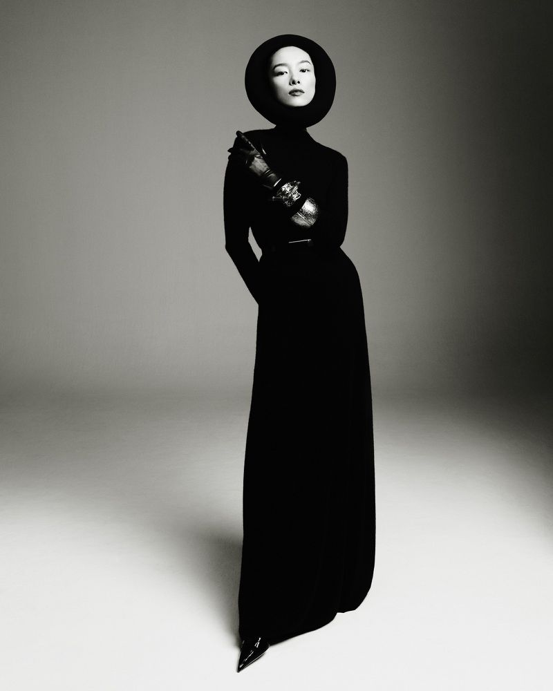 A stylish woman with pale skin and a black hat and dress stands in a photo studio in a high-fashion pose against a neutral backdrop.