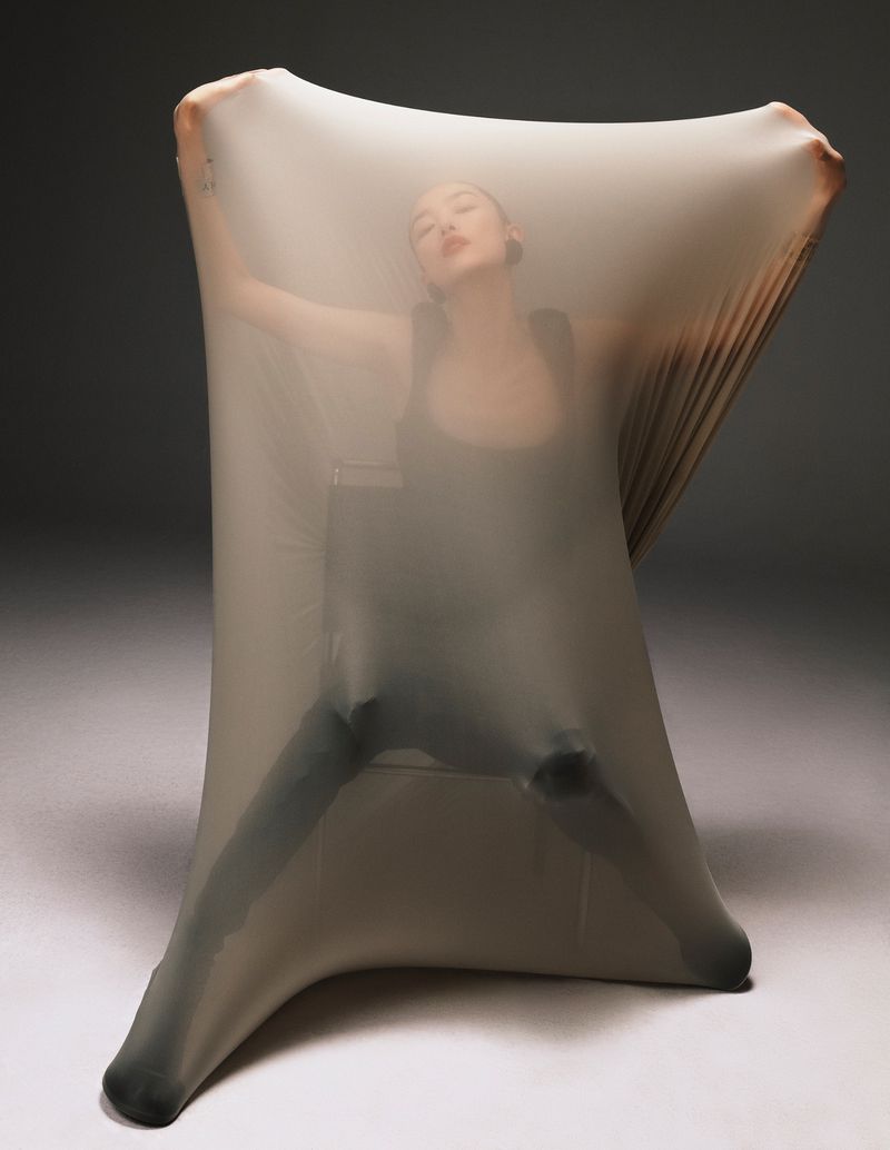 Artistic studio portrait of a female figure enveloped in stretched fabric against a neutral background, showcasing creative fashion and design.