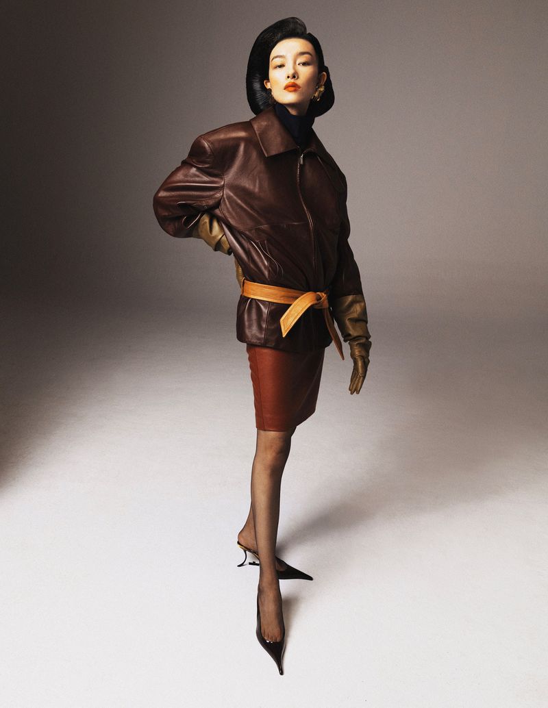 Elegant woman poses in a studio wearing a brown leather jacket, matching skirt, black stockings, and black high heels against gray background.
