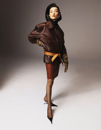 Elegant woman poses in a studio wearing a brown leather jacket, matching skirt, black stockings, and black high heels against gray background.