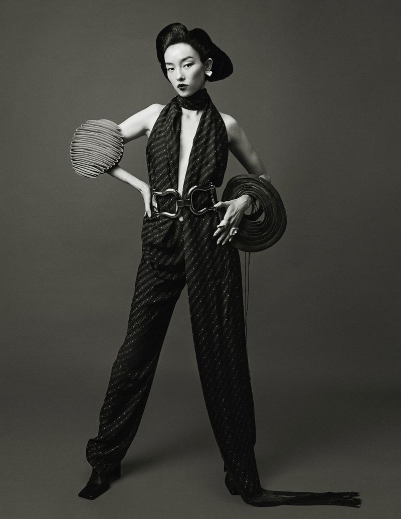 A stylish Asian model strikes a confident pose in a studio setting, showcasing a black patterned jumpsuit and sculptural hat.
