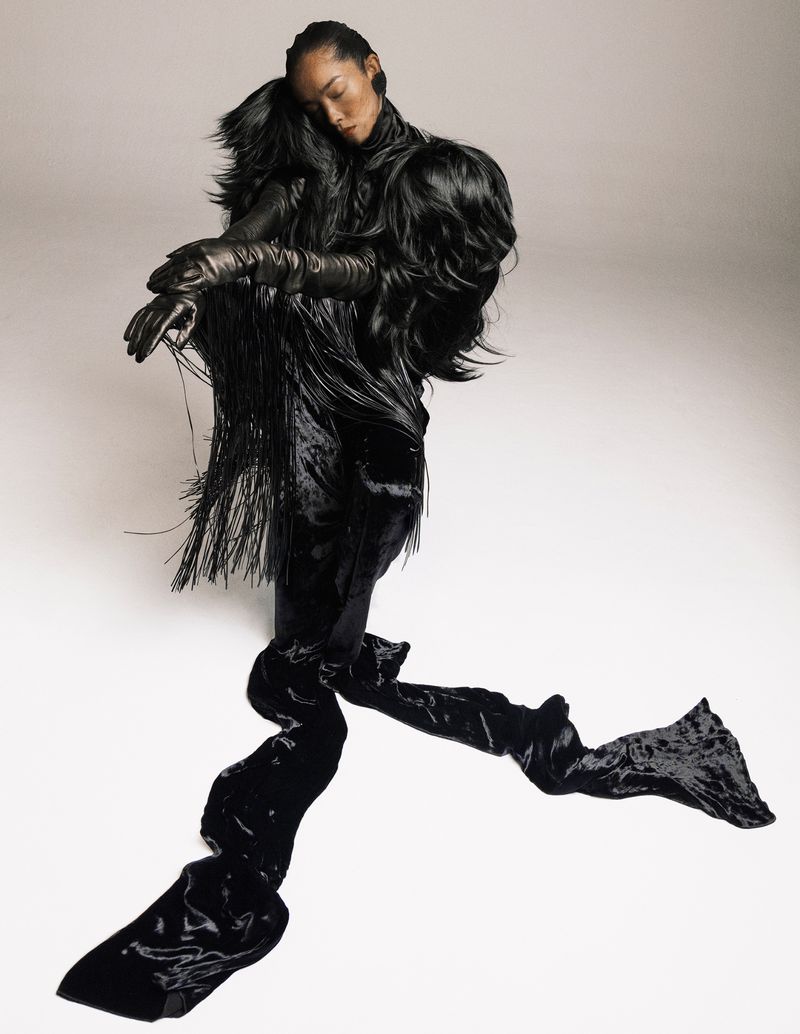 Elegant woman in avant-garde black gown with fringe detail, posing in a bright photo studio. High-fashion editorial shot.
