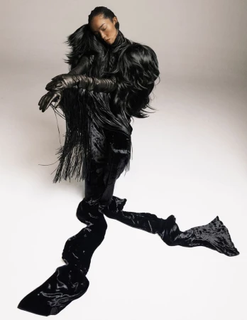 Elegant woman in avant-garde black gown with fringe detail, posing in a bright photo studio. High-fashion editorial shot.