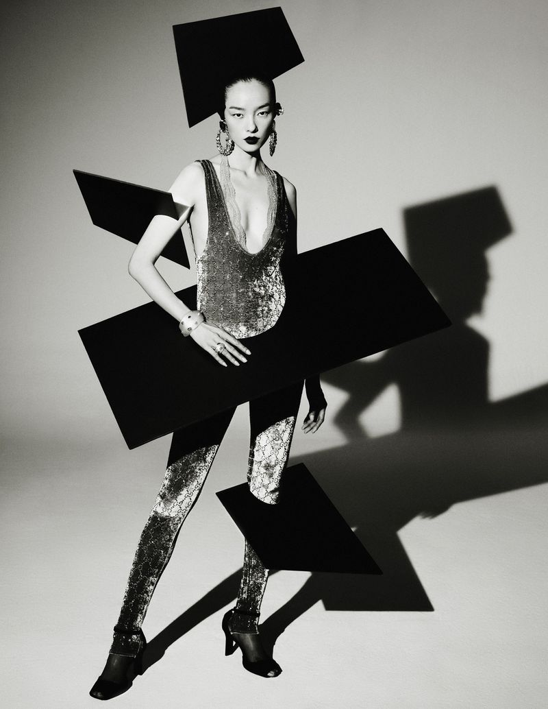 Monochrome fashion shot: Asian woman in sequined jumpsuit poses with geometric black shapes in a studio setting with theatrical lighting.