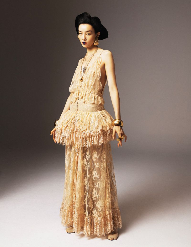 Elegant woman poses in a beige lace gown with gold jewelry against a neutral backdrop in a fashion photography studio setting.