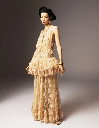 Elegant woman poses in a beige lace gown with gold jewelry against a neutral backdrop in a fashion photography studio setting.