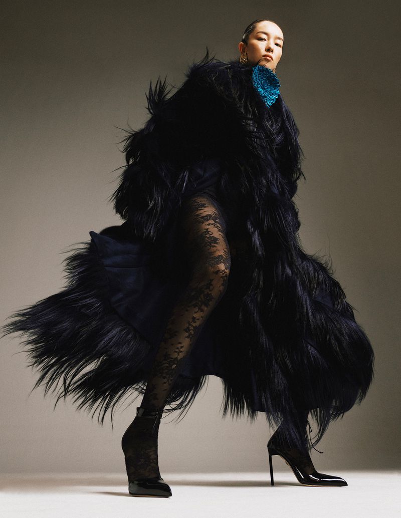 A woman poses dramatically in a long, dark, shaggy fur coat, black hosiery, and heels against a neutral studio backdrop, fashion forward.