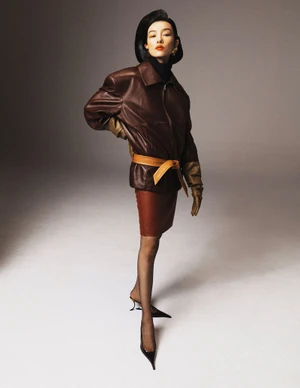 Model FeiFei Sun posing in an oversized brown leather jacket cinched with a tan belt, matching skirt, gloves, and pointed heels.