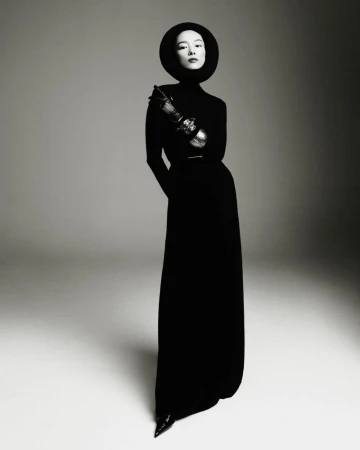 Model FeiFei Sun wearing a black floor-length dress and circular headpiece, posing against a neutral grey studio backdrop.