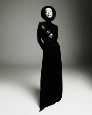 Model FeiFei Sun wearing a black floor-length dress and circular headpiece, posing against a neutral grey studio backdrop.