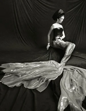 Model FeiFei Sun seated in a strapless corset and flowing translucent mermaid tail gown against a dark draped backdrop.