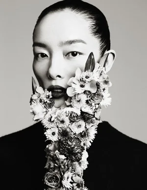 Black and white portrait of FeiFei Sun with a sculptural floral arrangement covering her jaw and neck against a studio backdrop.