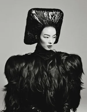 Model Fei Fei Sun wearing an avant-garde black sculpted square headdress and voluminous textured cape against a neutral studio backdrop.