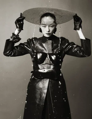 FeiFei Sun wears a black snakeskin coat and leather gloves while holding a dripping glass disc overhead in monochrome studio portrait.