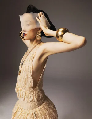 Fei Fei Sun posing in a cream ruffled lace dress, gold cuff bracelet, and translucent face veil against a grey studio backdrop.