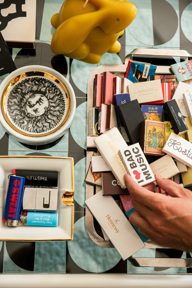 Overhead shot of a hand holding a matchbox amidst an array of decorative objects on a patterned surface, capturing a still life scene.