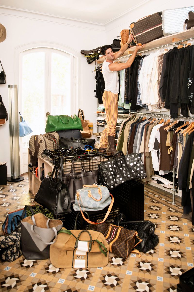 Man standing on a ladder in a walk-in closet placing a suitcase on a shelf among rows of hanging clothes, with many handbags on the floor.