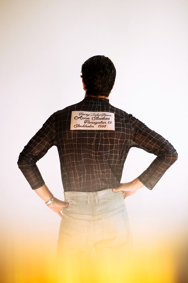 Back view of a person with short dark hair wearing a plaid shirt, light wash jeans, and a silver watch in a neutral indoor setting.