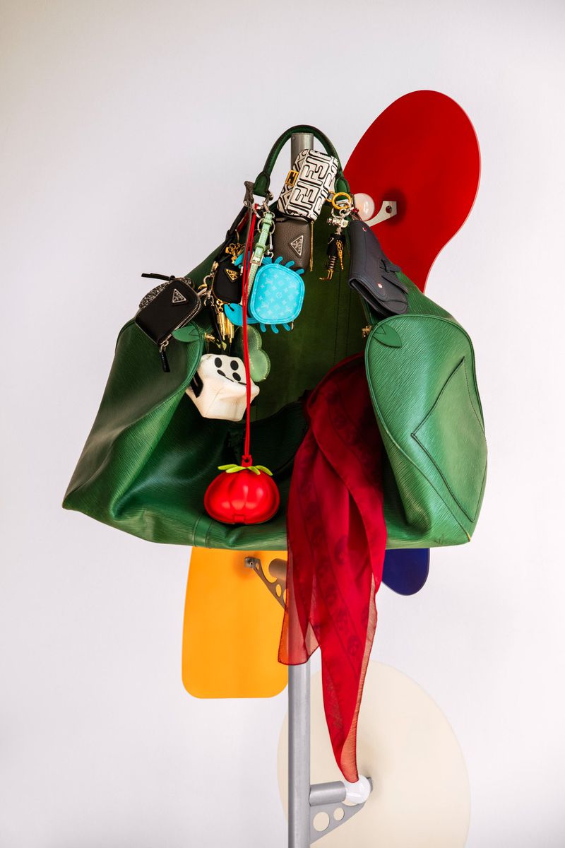 Green leather tote bag with charms, red scarf, and colorful board fragments hanging from a silver metal rack against a white wall.