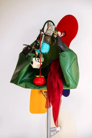 Green leather tote bag with charms, red scarf, and colorful board fragments hanging from a silver metal rack against a white wall.