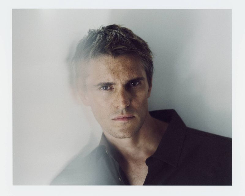 Close-up portrait of a man in a dark unbuttoned shirt looking at the camera with an artistic blur effect on the left.