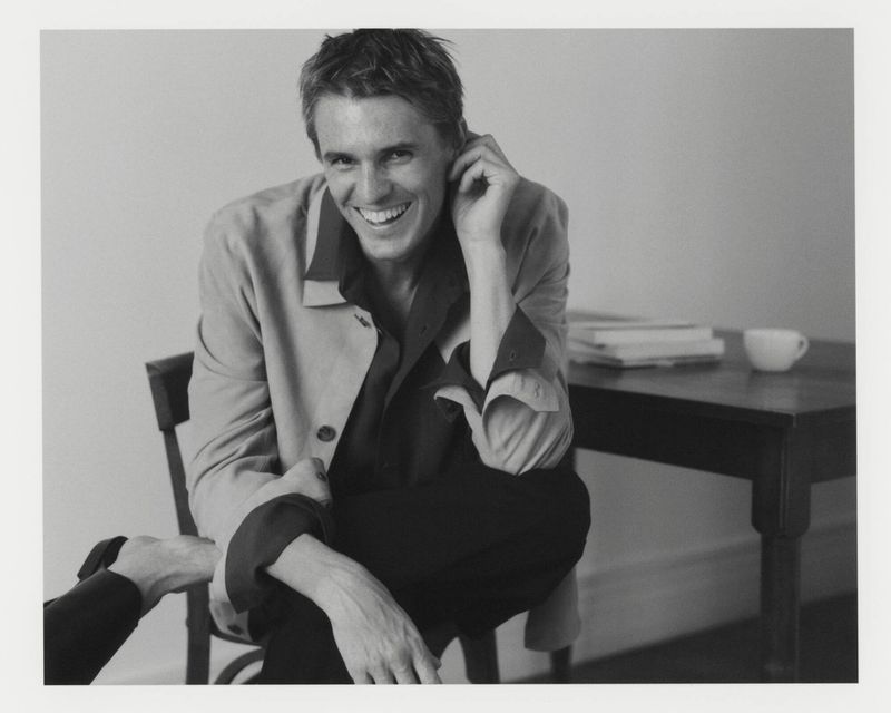 Adrien Dantou smiling while seated on a wooden chair beside a table in a monochrome Zara Minimal Trend AW25 menswear campaign image.