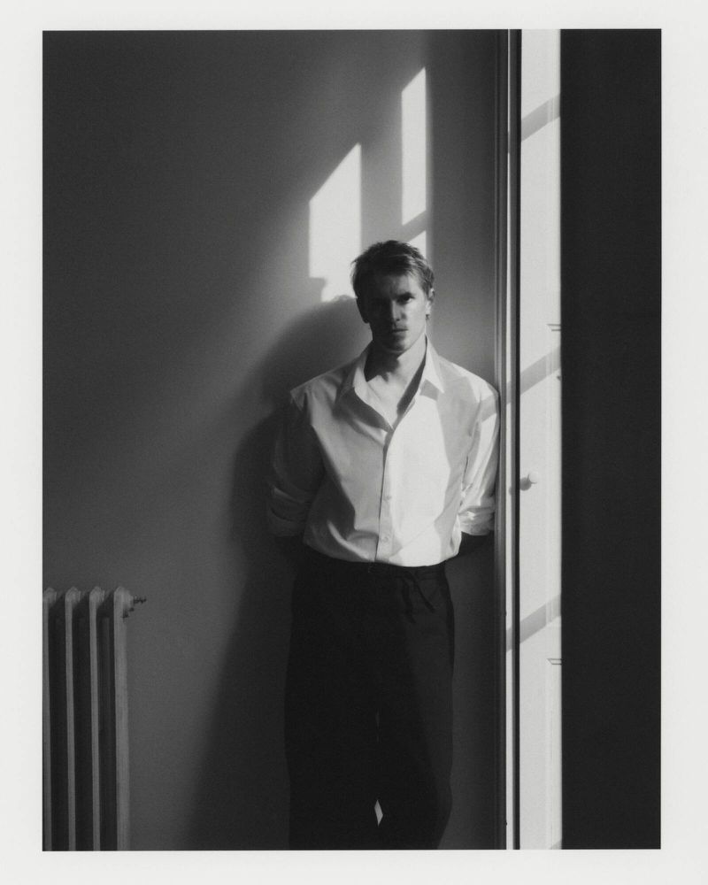 Monochrome portrait of a man in a white button-down shirt and dark trousers leaning against a sunlit window frame.