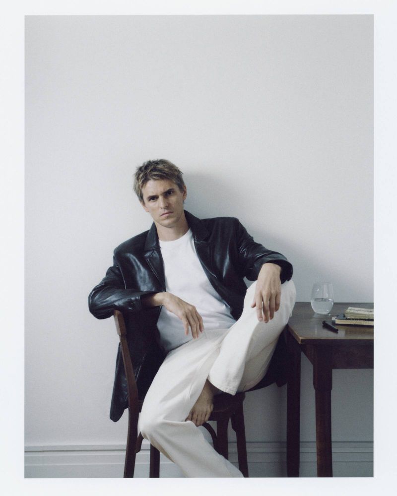 Man in a black leather jacket, white t-shirt, and white trousers seated on a wooden chair beside a small side table.