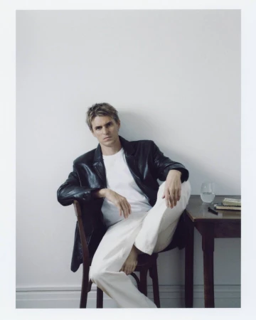 Man in a black leather jacket, white t-shirt, and white trousers seated on a wooden chair beside a small side table.