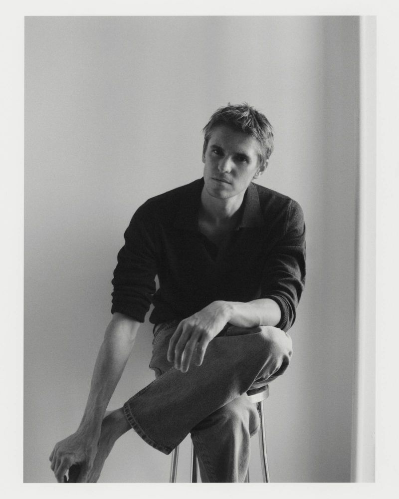 Man seated on a stool wearing a dark polo shirt and light trousers against a neutral background.