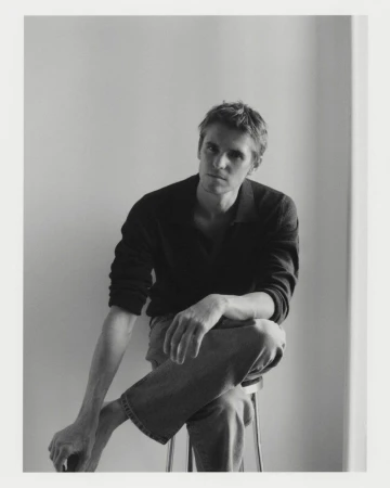 Man seated on a stool wearing a dark polo shirt and light trousers against a neutral background.