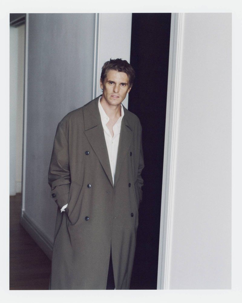 A man wearing an oversized olive double-breasted trench coat over a white open-collar shirt, standing in a doorway.