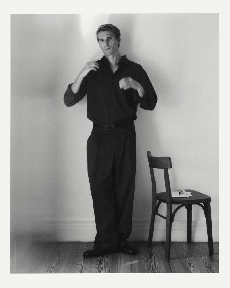 Monochrome portrait of an adult male adjusting his dark shirt, standing next to a wooden chair against a neutral wall in a studio setting.