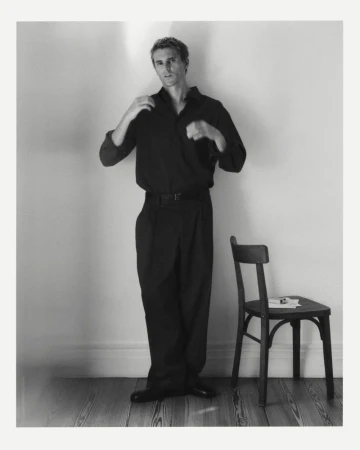 Monochrome portrait of an adult male adjusting his dark shirt, standing next to a wooden chair against a neutral wall in a studio setting.