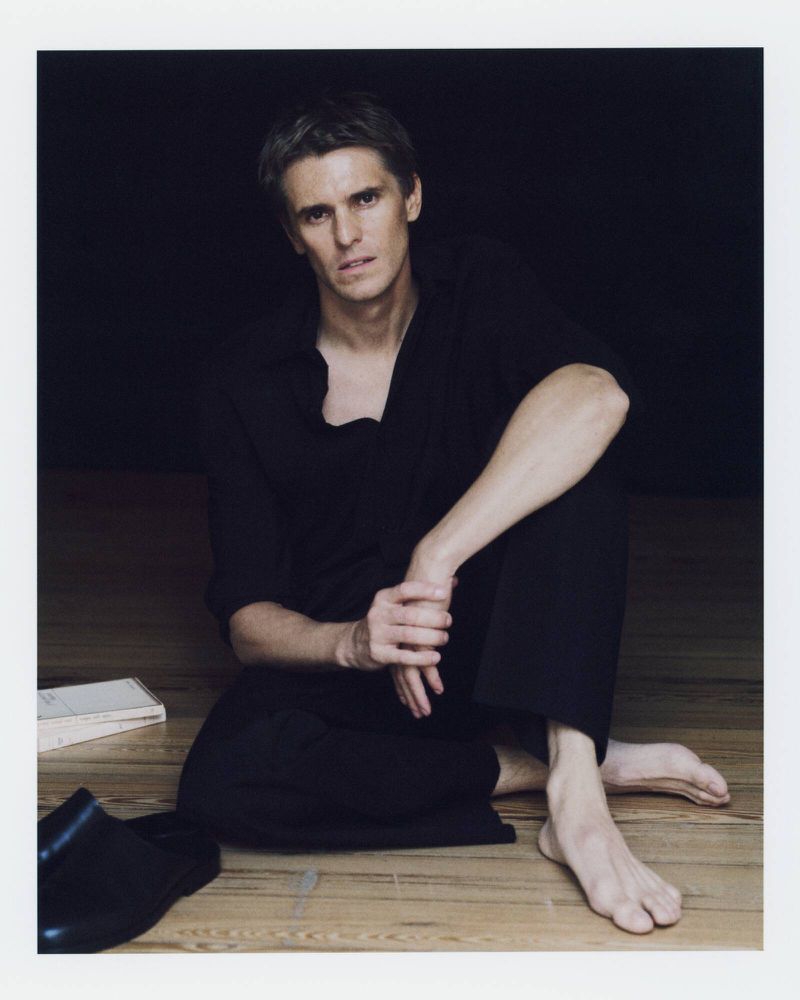 Adult male in black outfit sitting barefoot on a wooden floor next to a book and a shoe in front of a dark background.
