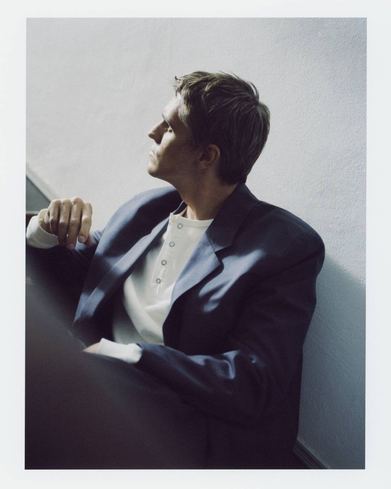 Indoor portrait of a man in a blazer sitting on stairs, looking away, with a neutral expression in soft lighting.