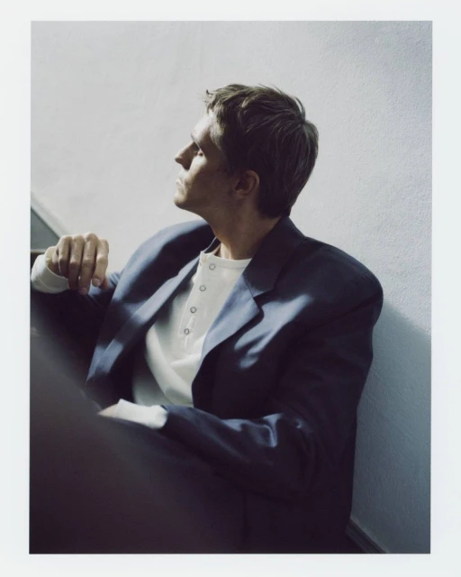 Indoor portrait of a man in a blazer sitting on stairs, looking away, with a neutral expression in soft lighting.