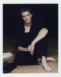 Adrien Dantou seated barefoot on a wooden floor wearing an all-black shirt and trousers for Zara.