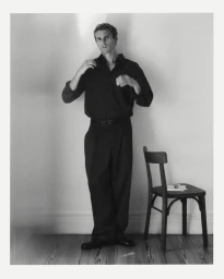 A man in a black button-down shirt and relaxed pleated trousers standing next to a vintage wooden chair against a plain wall.