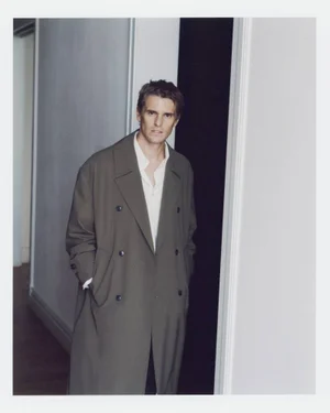 A man wearing an oversized olive double-breasted trench coat over a white open-collar shirt, standing in a doorway.