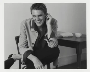 Adrien Dantou smiling while seated on a wooden chair beside a table in a monochrome Zara Minimal Trend AW25 menswear campaign image.