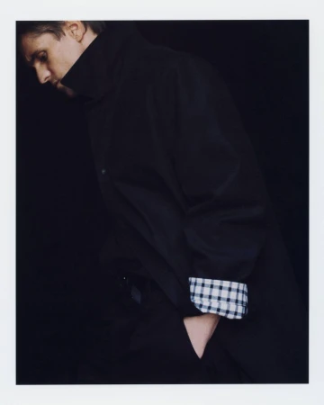 Man in side profile wearing a black high-collar coat with a gingham check cuff against a black background.